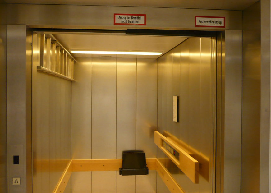 Bild 1 Feuerwehraufzug. An der linken Aufzugswand ist eine Leiter angebracht, in der hinteren Ecke links ist die Ausstiegsluke zu erkennen, im Bedientableau ist ein Schlüsselschalter (rote Aufschrift „Feuerwehr“) angebracht, welcher die Türen auch bei blockierter Lichtschranke (Rauch) schließen lässt.